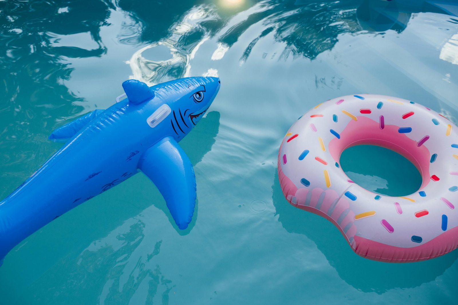 Brightly colored inflatable shark and donut floats in a sunlit swimming pool.